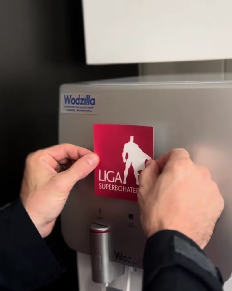 Wolontariusz Ligi Superbohaterów nakleja logo fundacji na nablatowy dystrybutor wody Wodzilla